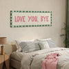 A framed canvas art print with the text LOVE YOU BYE in pink lettering on a white background, displayed above a bed.