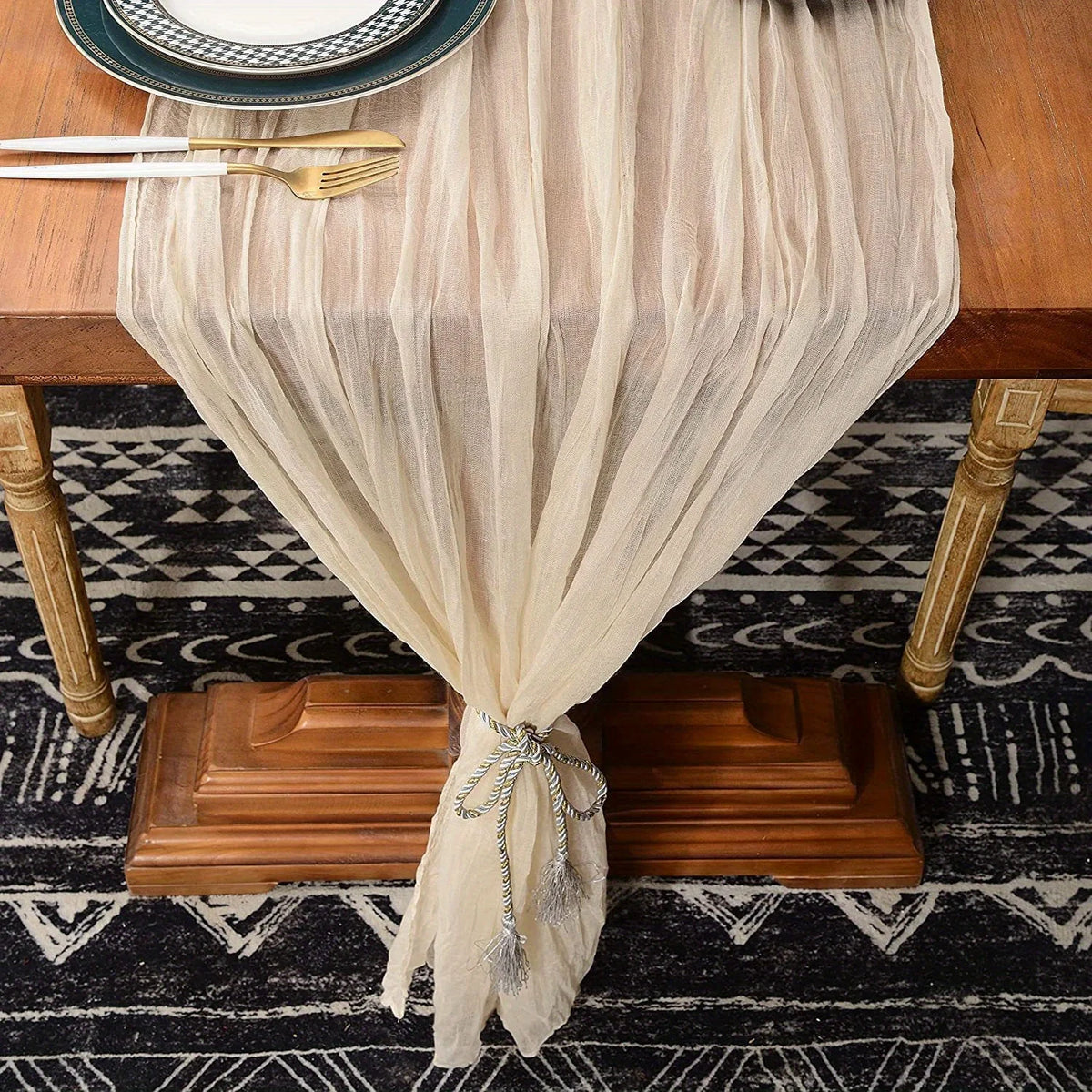 A beige cheesecloth table runner draped over a wooden table, featuring gold accents and fringed ties.