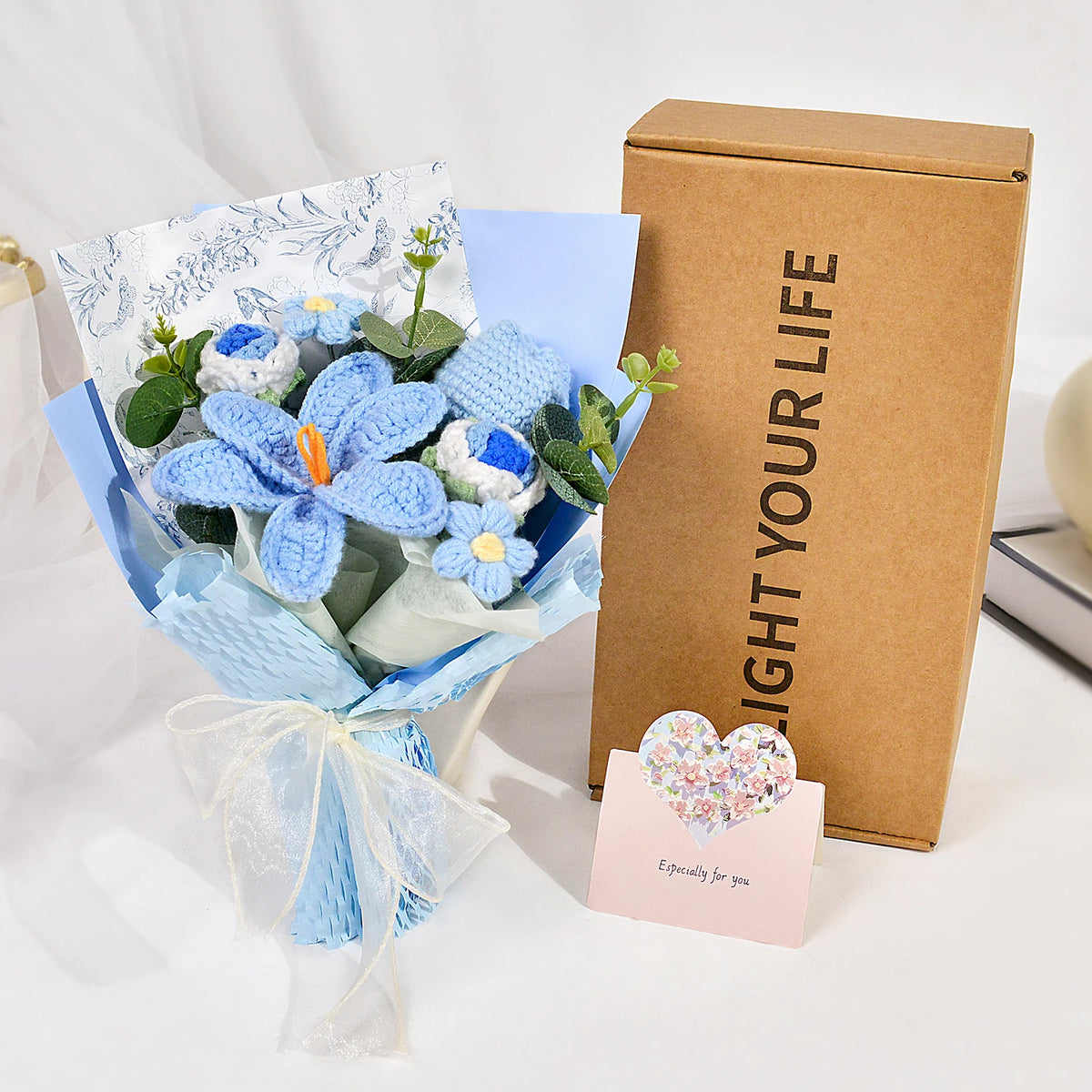 A bouquet of handmade crochet blue tulips wrapped in light blue paper next to a brown box labeled Light Your Life