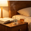A gourd-shaped glass incense burner with an ash catcher emitting smoke on a wooden bedside table.