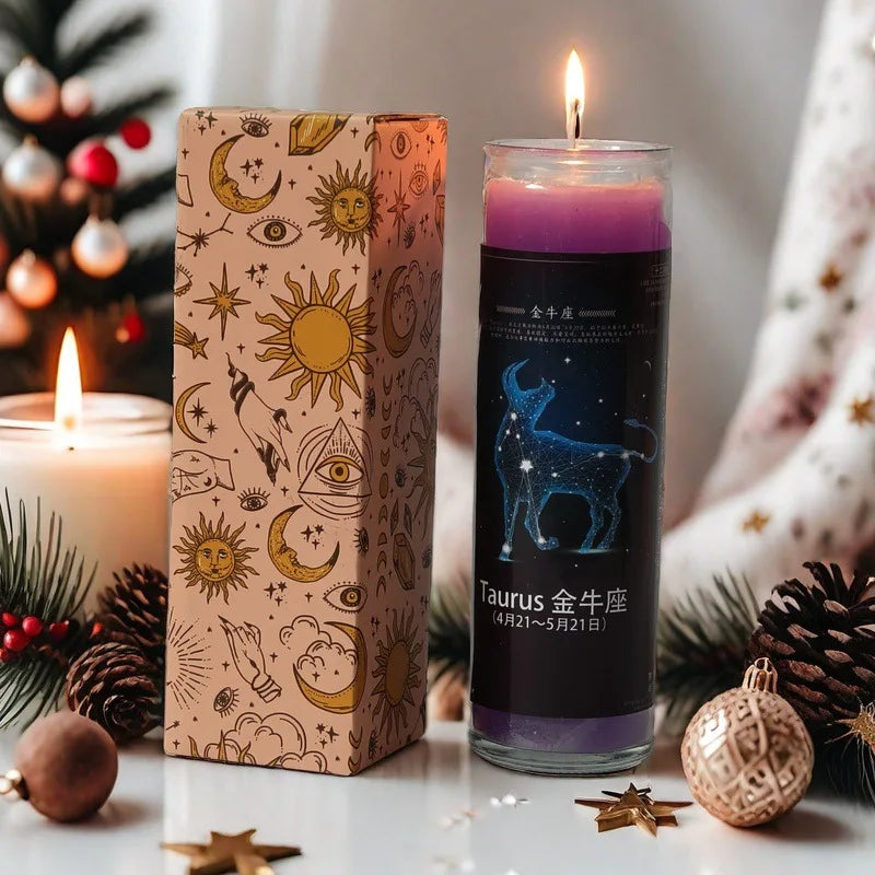 A glass candle in a rectangular box featuring a Taurus zodiac theme, set among pine branches and decorative ornaments.
