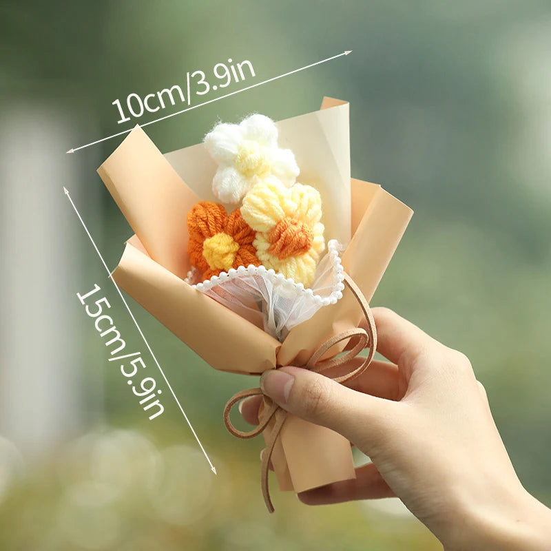A hand holds a bouquet of handmade crochet flowers in light orange and yellow tones, wrapped in kraft paper.