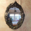 A dark, leafy wreath mirror framed in wood hangs on a wall, featuring an amulet.