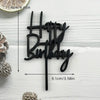 Black acrylic birthday cake topper with the words Happy Birthday on a white wooden background.
