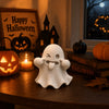 A white kawaii ghost statue with a tongue sticking out sits on a wooden table next to Halloween decor.
