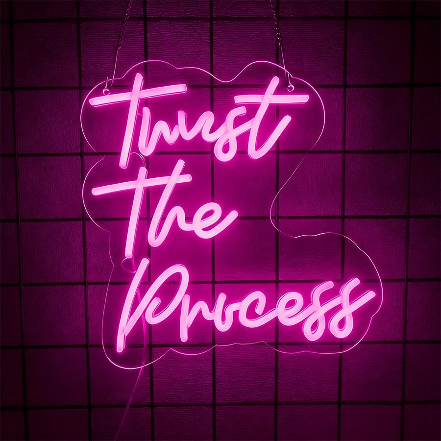 Pink neon sign spelling out Trust The Process on a dark purple grid background.
