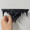 A hand holds a black melted skull wall shelf with dripping features against a white background.