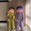 A pair of fuzzy monster onesie pajamas, one green and one purple, standing in a modern room.