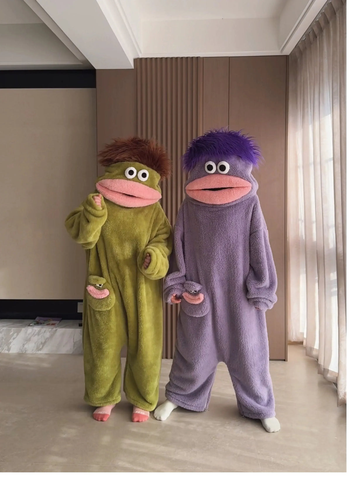 A pair of fuzzy monster onesie pajamas, one green and one purple, standing in a modern room.