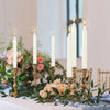 A display of ivory white drip-free stick candles in a golden holder arranged on a white surface.