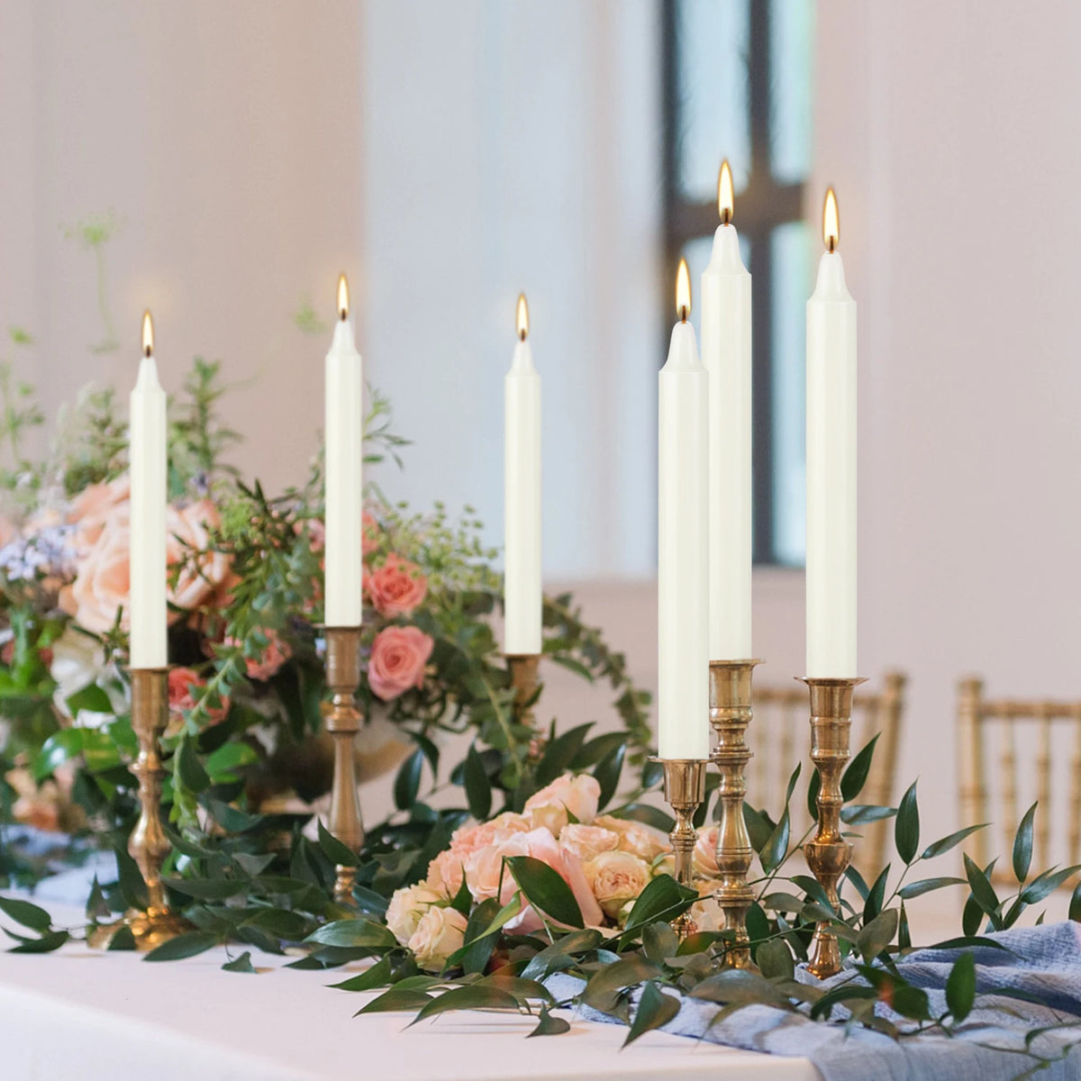 A display of ivory white drip-free stick candles in a golden holder arranged on a white surface.