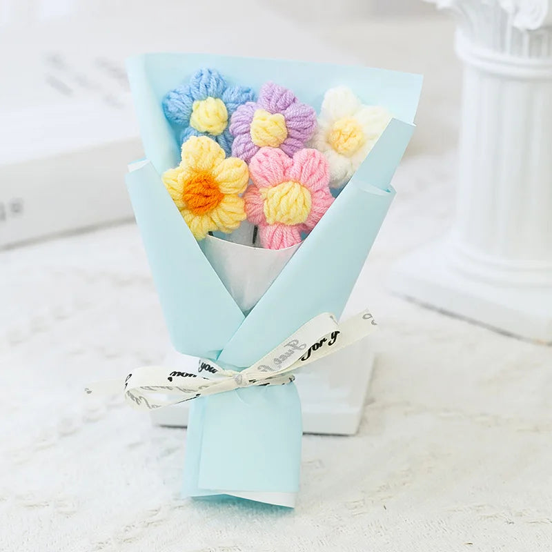 A bouquet of handmade crochet flowers in pastel blue, yellow, and purple arranged in a folded paper presentation.