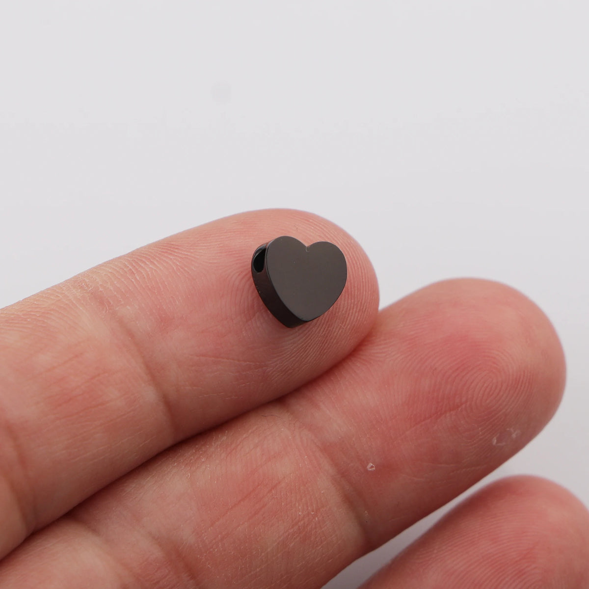 A hand holds a small, heart-shaped, dark gray stainless steel bead charm.