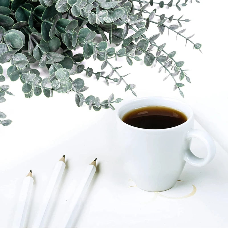 A white mug of coffee sits next to green artificial eucalyptus leaves on a white surface with pencils.