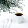A white mug of coffee sits next to green artificial eucalyptus leaves on a white surface with pencils.