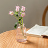 A clear glass vase holds pink roses on a wooden table next to an open book.