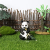 A realistic panda figurine sitting on green grass in front of a wooden fence and tropical plants.