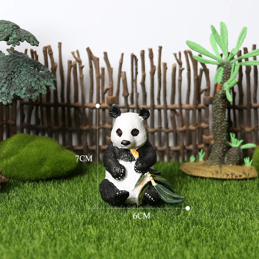 A realistic panda figurine sitting on green grass in front of a wooden fence and tropical plants.