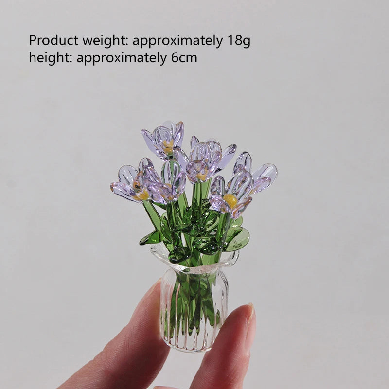 A hand holds a small bouquet of purple glass flowers in a clear glass vase.