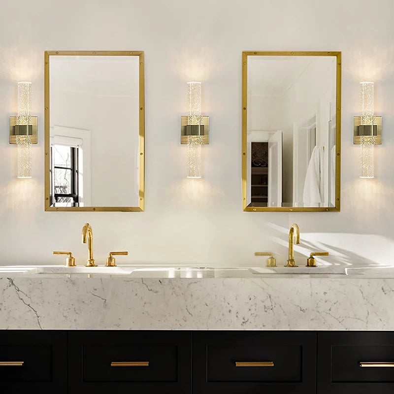 A bathroom vanity featuring a marble countertop, gold faucets, and two rectangular mirrors illuminated by modern bubble cryst