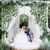 A bride and groom stand under a white draped archway decorated with green ivy garlands in an outdoor setting.