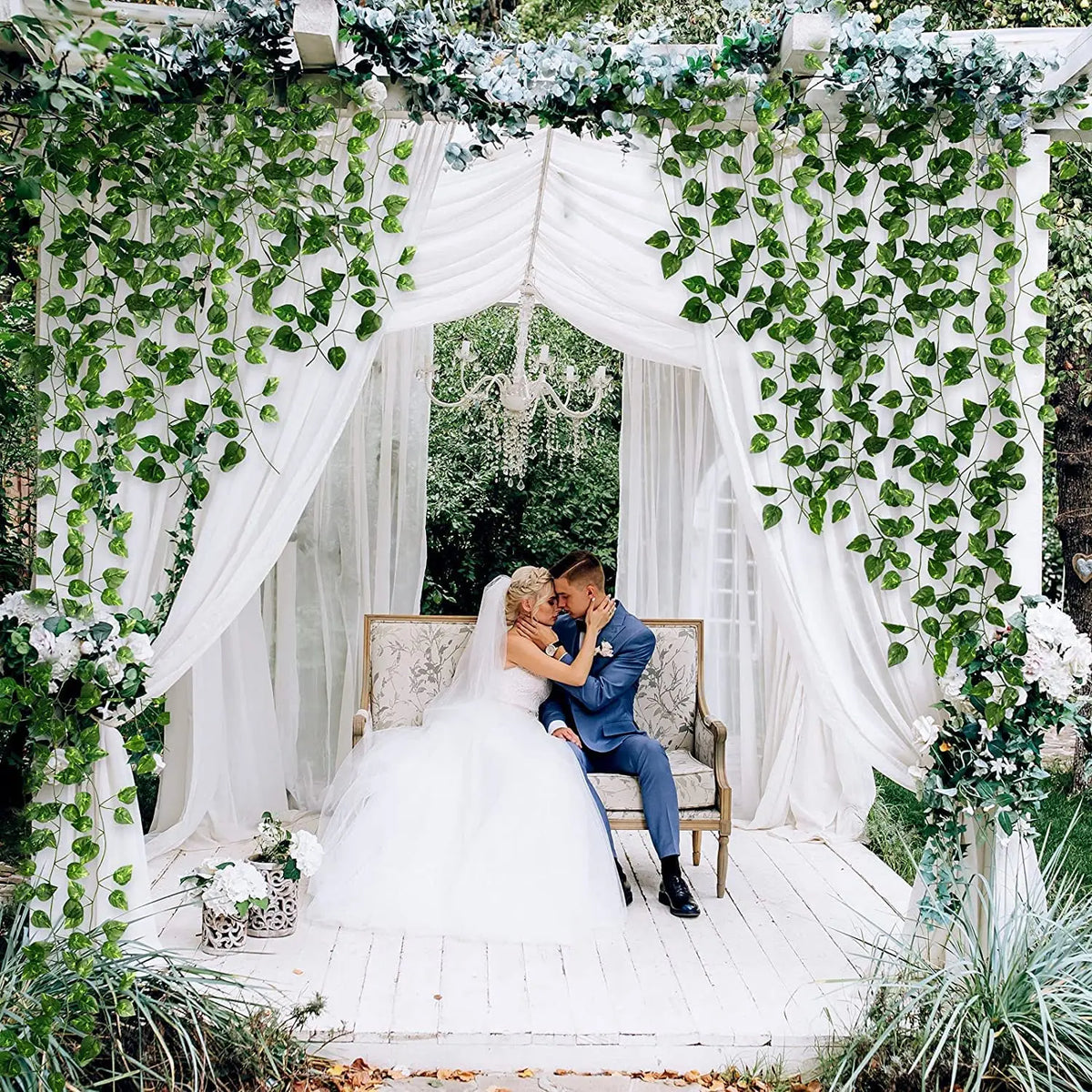 A bride and groom stand under a white draped archway decorated with green ivy garlands in an outdoor setting.