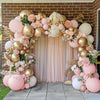 A pink and gold balloon garland arch decoration featuring roses, greenery, and balloons against a brick wall.