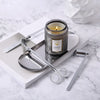 A glass jar candle sits on a silver tray next to a pair of silver scissors and a white notebook on a marble surface.