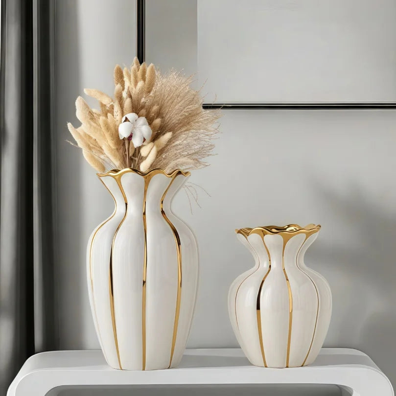 A white ceramic flower vase with gold accents sits on a shelf next to a smaller matching vase.