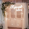 A neon sign spelling out All you need is love is displayed on a wooden cabinet decorated with white floral greenery and sheer