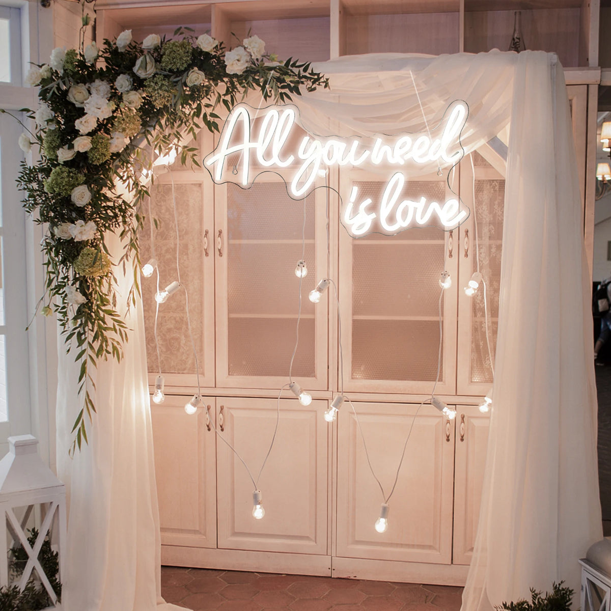 A neon sign spelling out All you need is love is displayed on a wooden cabinet decorated with white floral greenery and sheer
