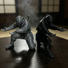Two dark ninja figures in gray and black are shown standing on a wooden desk with smoke effects.