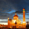 A golden Islamic mosque building at dusk with a prominent minaret and dome under a dramatic sky.