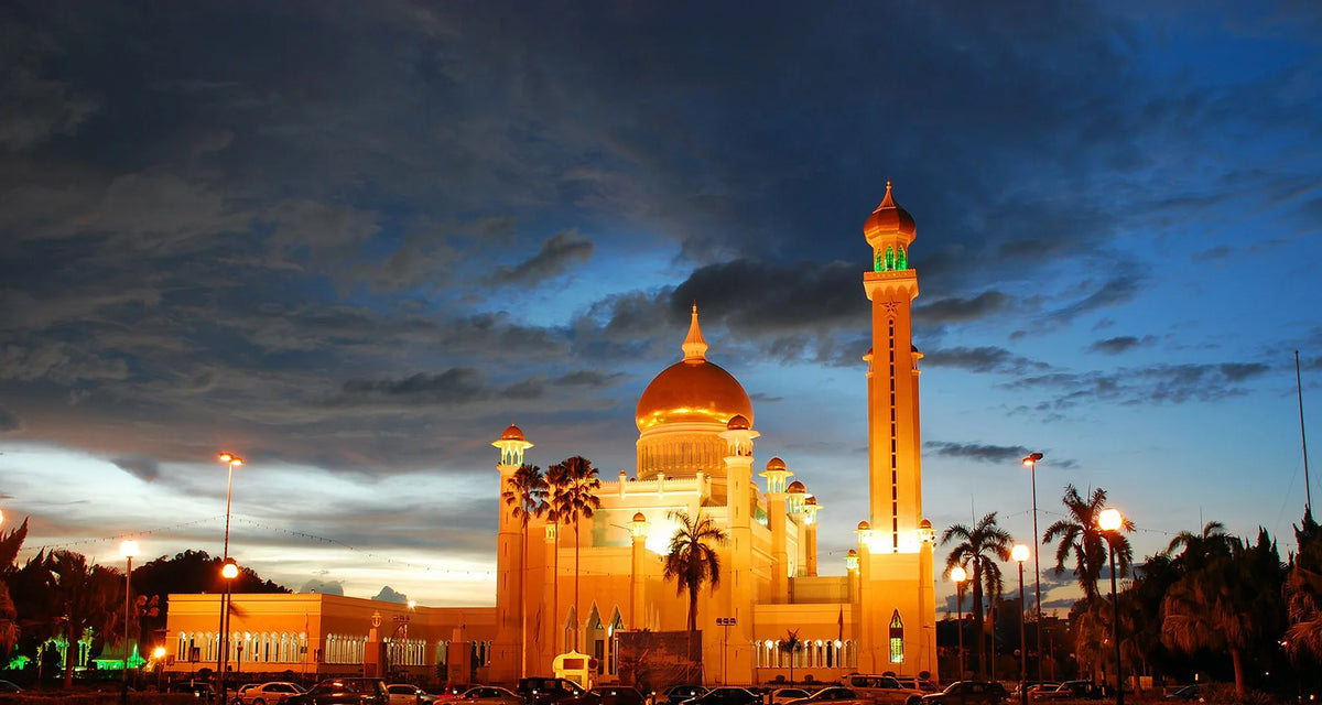 A golden Islamic mosque building at dusk with a prominent minaret and dome under a dramatic sky.