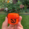An orange miniature pumpkin Halloween bucket with a carved face is held in a hand against a blurred green outdoor background.