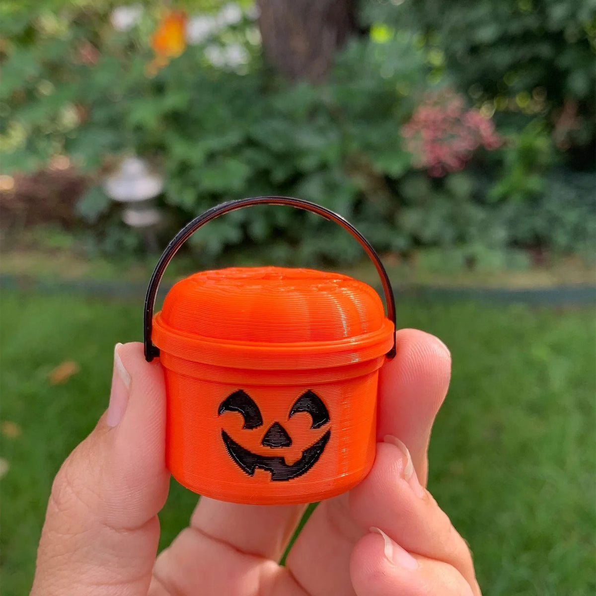 An orange miniature pumpkin Halloween bucket with a carved face is held in a hand against a blurred green outdoor background.