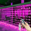 A hand holds a remote control for remote control LED curtain fairy lights illuminating a building facade with pink light.