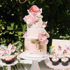 A tiered pink cake decorated with floral elements and fairy toppers, displayed outdoors.