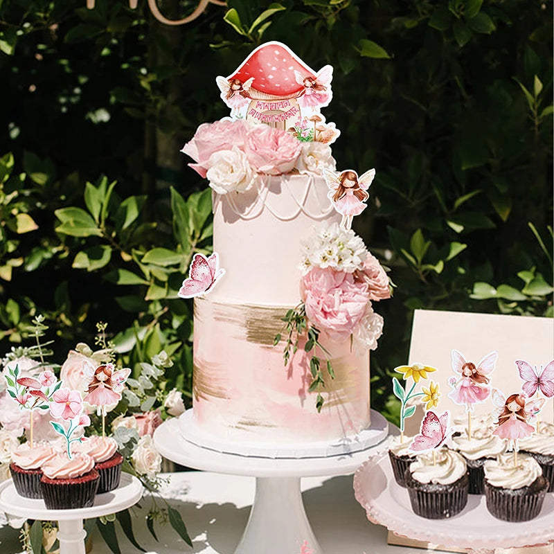 A tiered pink cake decorated with floral elements and fairy toppers, displayed outdoors.
