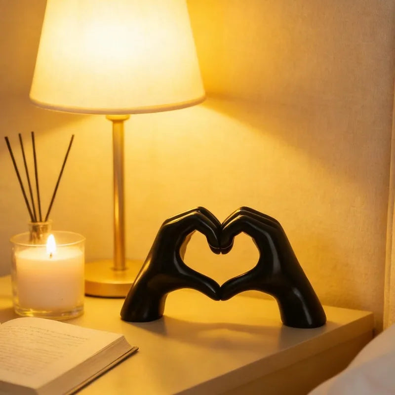 A black heart hand sculpture is displayed on a bedside table next to a lamp and a candle.