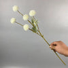Una mano sostiene un tallo de flores artificiales de margarita blanca contra un fondo gris.