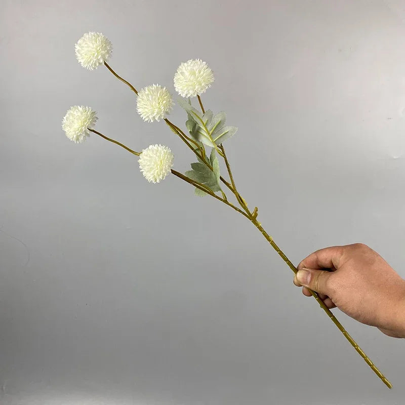 Una mano sostiene un tallo de flores artificiales de margarita blanca contra un fondo gris.