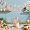 Pink tulle bow garland strung across a light blue wall above wooden shelves and a dessert display.