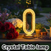 An oval crystal LED table lamp with a glowing numerical zero is displayed on a wooden surface next to flowers.