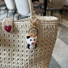A handmade crochet cow keychain with a white and black pig face hanging from a beige woven tote bag.