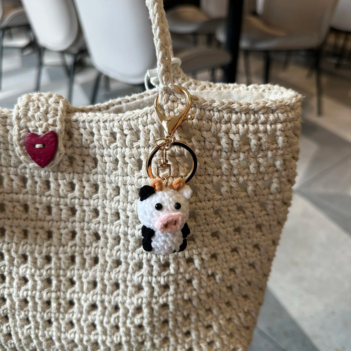 A handmade crochet cow keychain with a white and black pig face hanging from a beige woven tote bag.
