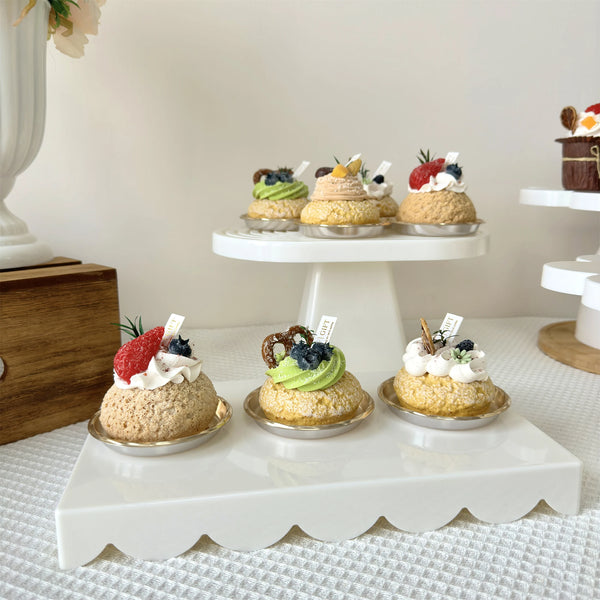 A white display stand holds two small cupcakes with cream and fruit toppings on a white surface.