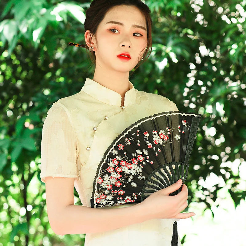 A woman in a cream-colored top holds an elegant black folding fan decorated with red and white plum blossoms.