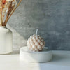 A white ceramic candle holder with a spherical cluster of white wax bubbles on a light surface.