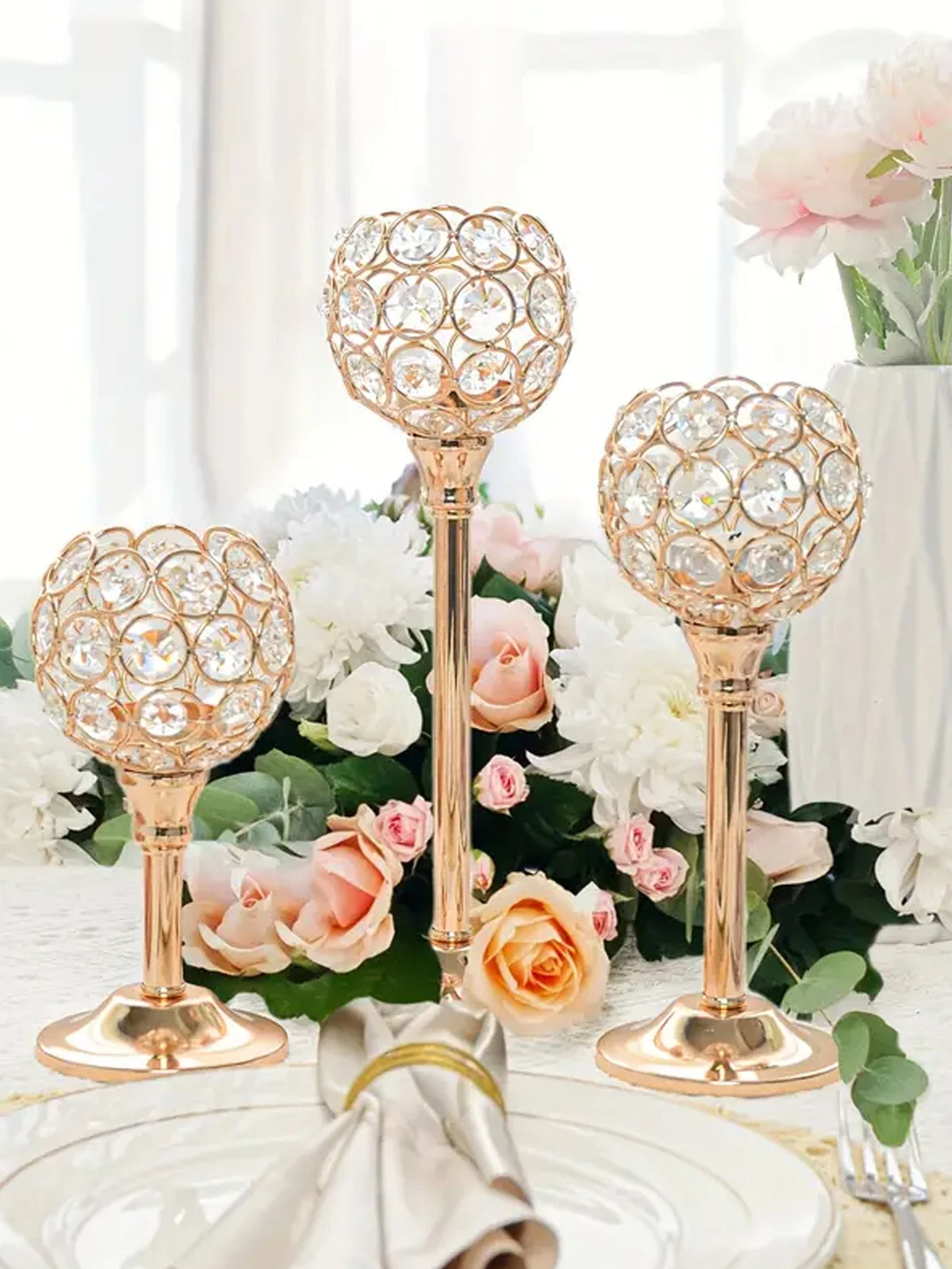 A set of golden crystal candle holders with floral designs arranged on a white table surrounded by pink and white roses.